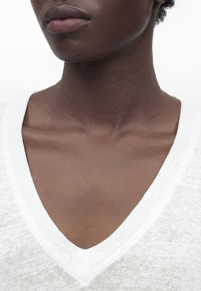 Close-up of a person’s neck, lower face, and collarbone wearing a white V-neck garment against a plain background.