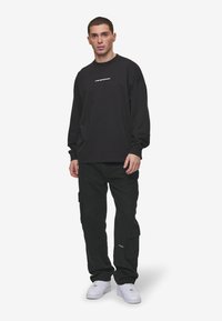 Camisa negra de manga larga con logo blanco, combinada con pantalones cargo negros con bolsillos y zapatillas blancas. Look sencillo y casual.