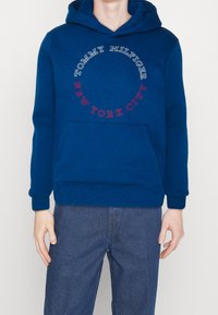 Modrá mikina s kapucí a přední kapsou, s žebrovanými manžetami. Obsahuje text „TOMMY HILFIGER“ a „NEW YORK CITY“ ve formě kruhové grafiky. Džíny na pozadí.