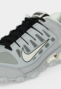 Γκρι και μαύρο αθλητικό παπούτσι Nike με λευκό λογότυπο swoosh, μαύρα κορδόνια και λευκό μαξιλαράκι στον γιακά, εμφανίζεται από πλάγια γωνία.