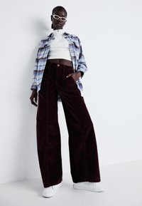 Bourgondische corduroy broek met wijde pijpen, gedragen met een witte crop top en een blauwe geruite blouse, met een ontspannen pasvorm en zijzakken. Witte sneakers.