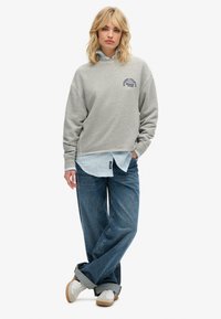 Sudadera gris con un logo redondo, llevada sobre una camisa de botones azul claro. Combinada con jeans holgados de color azul y zapatillas blancas.