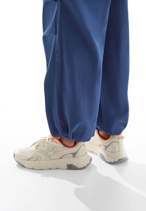 Baskets crème avec des semelles texturées, des accents gris et des touches orange, accompagnées d'un pantalon bleu ample resserré aux chevilles.