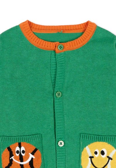 Cardigan vert en tricot avec bordure orange, boutons sur le devant et poches ornées de visages souriants de balle de basket et de balle de tennis.