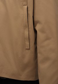 Nærbillede af et ærme på en beige jakke og en sidelomme med en lodret slids, der viser glat stof og fine syningsdetaljer.