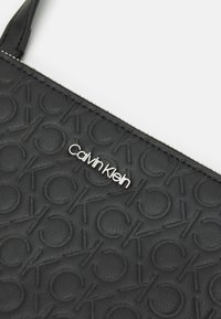 Černá kožená taška s embosovaným vzorem loga, se stříbrným emblémem "Calvin Klein" a zipovým uzávěrem podél horní hrany.