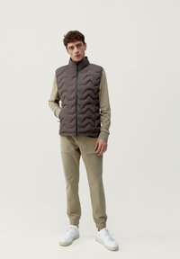 Donkerbruine puffervest met een quilted zigzagpatroon, gecombineerd met een beige shirt met lange mouwen en bijpassende taps toelopende broek, witte sneakers.