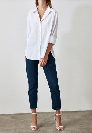 Femme portant une chemise blanche ample à boutons, un jean court bleu foncé et des talons blancs ouverts à l'avant, debout sur un sol gris contre un mur blanc.