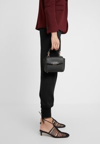 Zwarte, gestructureerde handtas met een bovenhandvat en gouden hardware, gecombineerd met zwarte broek en zwarte sandalen met bandjes.