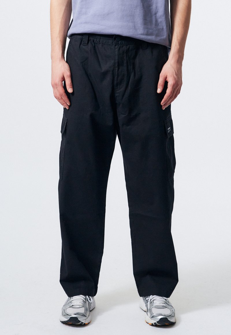 Černé cargo kalhoty s volným střihem, elastickým pasem a bočními kapsami, doplněné o šedé sportovní boty. Hladká textura a jednoduchý design.