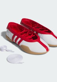 Witte en rode binnenshuis schoenen met zachte zool, rode veters en twee extra paar witte veters ernaast op een lichte achtergrond.