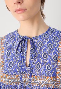 Blå mønstret bluse med blomster- og geometriske design, med en nøkkelhulls halsbånd og teksturert stoff. Innslag av oransje og hvitt.