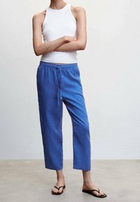 Blauwe linnen broek met een elastische tailleband en trekkoord. Heeft een ontspannen pasvorm en cropped lengte, gecombineerd met een wit tanktop en sandalen.