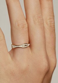 Anillo en tono dorado con un diseño ondulado, que presenta el texto grabado "LUNA" y un pequeño acento de gema. Textura suave, estilo minimalista.