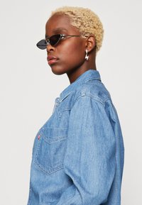 Lichtblauwe denim overhemd met een button-down kraag, voorzien van een borstzak en een rood label. Zwarte zonnebril en een bliksemschichtoorbel als accent.