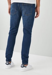 Jeans de mezclilla azul oscuro con un corte slim, que cuentan con dos bolsillos traseros y detalles sutiles de costuras, combinados con zapatillas blancas.