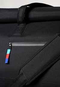 Černá taška na rolovací uzávěr z texturovaného materiálu, s horním popruhem, zipovou kapsou a barevným akcentem na táhlu zipu.