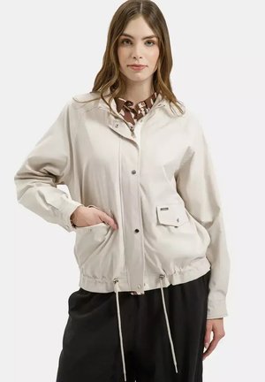 Młoda kobieta w jasnobeżowej kurtce zapinanej na zamek z sznurkami, dużymi przednimi kieszeniami oraz czarnych spodniach o szerokich nogawkach, stojąca na tle białego tła.