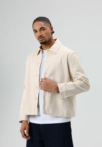 Beige jas met een knoopsluiting aan de voorkant, een spreide kraag en lange mouwen. Draagt over een wit T-shirt, van een gestructureerde stof en met een minimalistisch ontwerp.