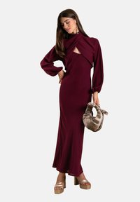 Bordeauxfarbenes Langarmkleid mit Ausschnitt und Ballonärmeln, kombiniert mit einer Handtasche mit goldenen Akzenten und Plattformschuhen. Glattes Material.