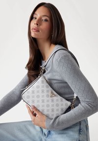 Witte handtas met een grijs logo patroon, metalen hardware en een triangulaire accent. Draagcrossbody, gecombineerd met een grijze textuurtrui.