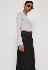 Világoslila, hosszúujjú cropped top, textúrált mintával és fodros mandzsettával, magas derékú fekete szoknyával párosítva, laza testtartásban.