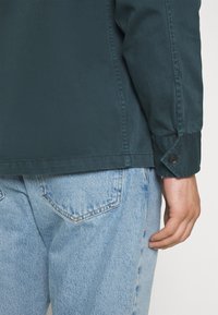 Person trägt ein dunkelgrünes, langärmliges Hemd mit Knöpfen und hellblaue Jeans aus Denim, die Hand locker an der Seite.