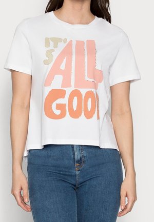 Žena ve bílé zkrácené tričko s nápisem "VŠECHNO JE DOBRÉ" v béžové, růžové a oranžové barvě, spárované s modrými džíny.