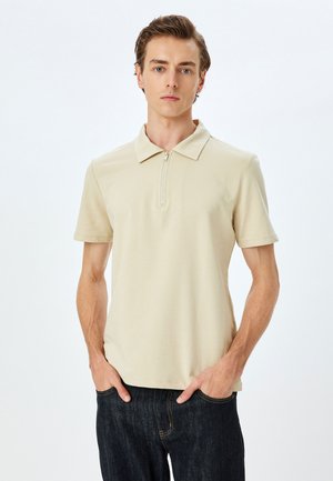 Camisa polo beige de manga corta con cuello de cremallera, hecha de tela suave. Combinada con jeans de mezclilla oscuros, llevada de manera casual con las manos en los bolsillos.