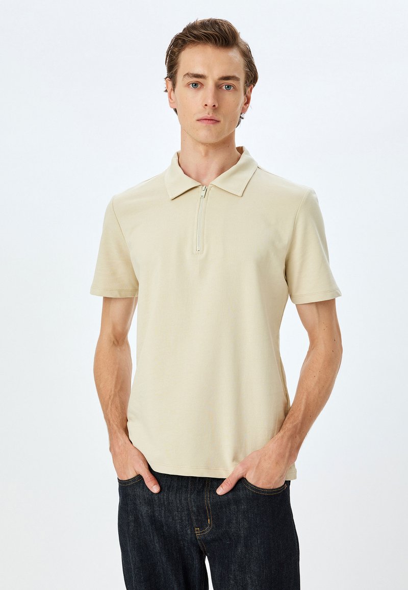 Beige polo-shirt med korte ærmer og lynlås krave, lavet af blødt stof. Båret med mørke denimjeans, brugt afslappet med hænderne i lommerne.