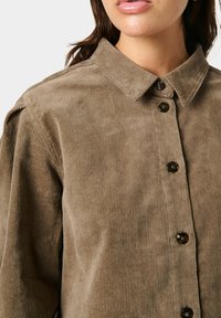 Khaki korduroy marškiniai su apykakle, priekiniais mygtukais ir siauru tekstūru. Tamsios, blizgios apdailos mygtukai prideda detalumo.