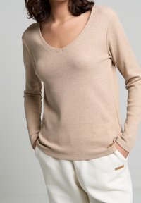 Top beige de manga larga con escote en V, textura acanalada y aberturas laterales, combinado con pantalones de crema claros con textura y detalles metálicos.