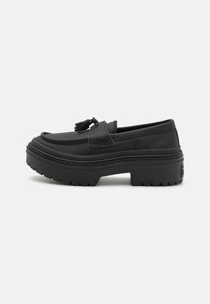 Mocasín de cuero negro con suela de plataforma gruesa y detalle de borla en la parte superior, mostrado de perfil lateral sobre fondo blanco.
