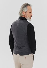 Szary kamizelka o teksturowanym wykończeniu, z regulowanymi guzikami z tyłu, noszona na czarnej koszuli z długim rękawem, zestawiona z jasnymi spodniami.