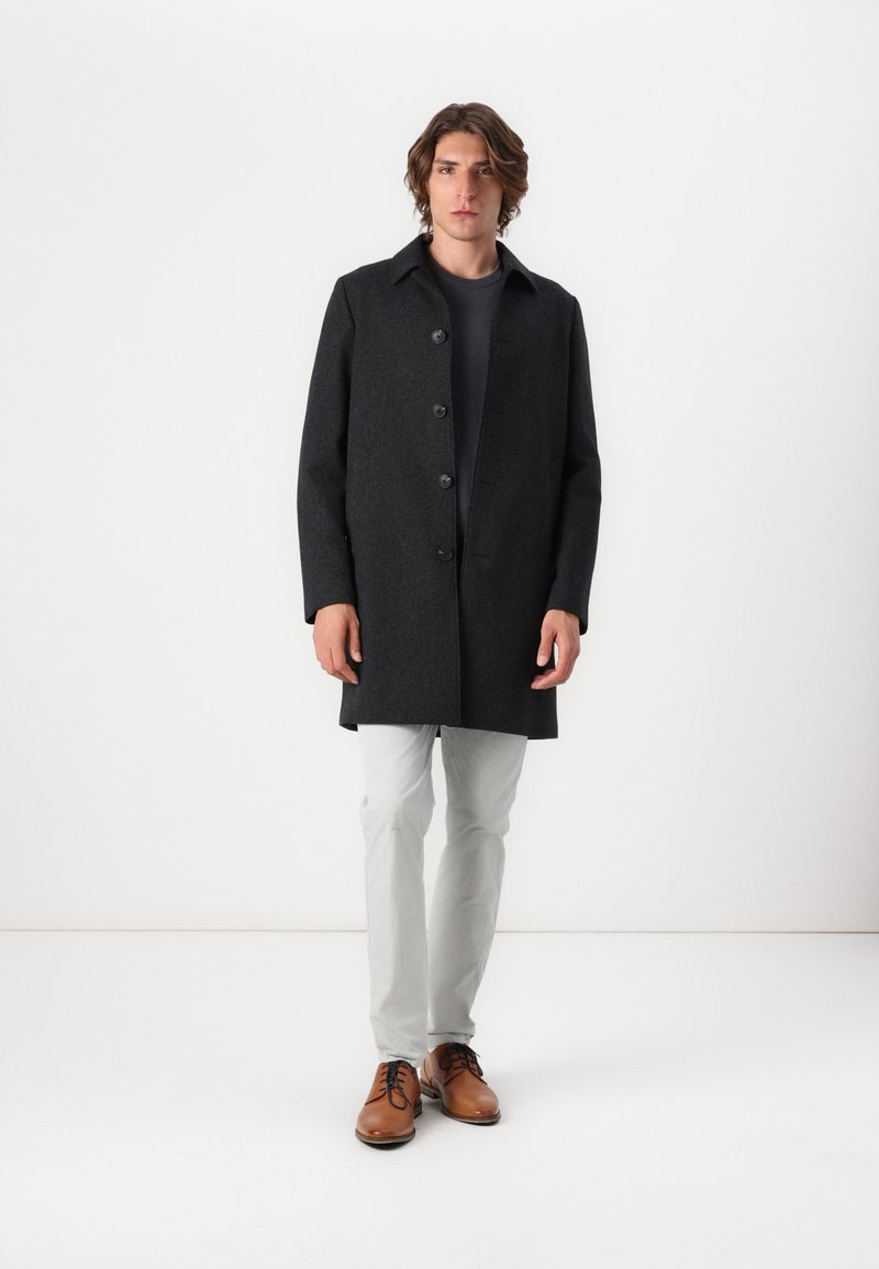 Mann in einem dunkelgrauen knopfartigen Mantel, schwarzem Hemd, hellgrauen Hosen und braunen Schuhen, der vor einem weißen Hintergrund steht.