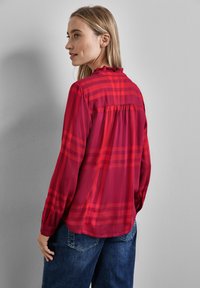 Blusa rossa a scacchi con scollatura arricciata, maniche lunghe e vestibilità rilassata. Presenta una piega sul retro e un tessuto morbido e leggero.