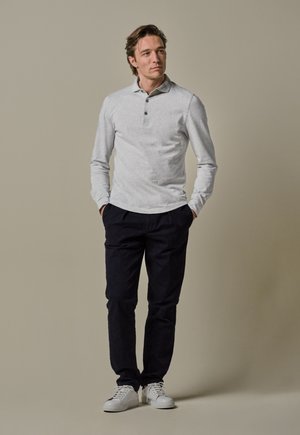 Uomo in piedi con le mani in tasca, che indossa una polo a maniche lunghe grigia chiara, pantaloni neri e sneakers bianche, su uno sfondo neutro.