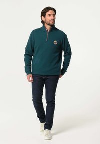 Groene rits fleece pullover met een staande kraag, geborduurd logo, gecombineerd met donkere jeans en witte sneakers. Glad textuur en een relaxte pasvorm.