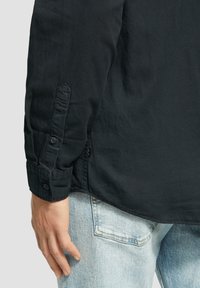 Camisa negra de botones con mangas enrolladas, que presenta una textura suave y un ligero brillo, combinada con jeans de mezclilla azul claro visibles en la cintura.