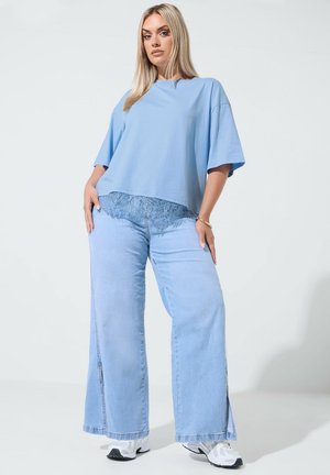 Blonde vrouw met een losse lichtblauwe top met kanten zoom, wijdvallende lichtblauwe spijkerbroek en witte sneakers, staand tegen een effen achtergrond.