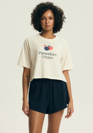 Kobieta o kręconych włosach ubrana w beżowy krótki t-shirt z grafiką pomidora i napisem, w połączeniu z czarnymi szortami na sznurku.