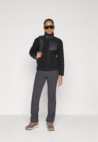 Persoon in een zwarte fleece jas, donkergrijze broek, wandelschoenen, een zonnebril en met een zwarte rugzakriem over één schouder.