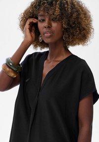 Czarny tunikowy top z dekoltem w serek i krótkimi rękawami, wykonany z lekkiego materiału. Dodatki to nałożone na siebie bransoletki i wzorzyste kolczyki.