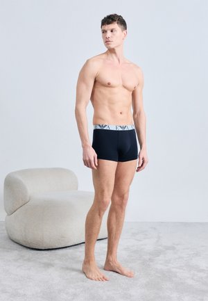 Mężczyzna bez koszulki stojący boso na wykładzinie, ubrany w czarne bokserki z białym pasem, obok zakrzywionego beżowego fotela.