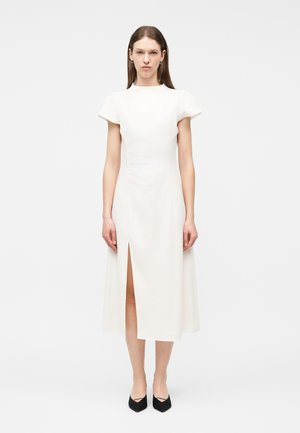 Γυναίκα που στέκεται μπροστά σε λευκό φόντο, φορώντας ένα λευκό φόρεμα μέσης μάκρους με κοντά μανίκια και μπροστινή σχισμή, συνδυασμένο με μαύρα μυτερά παπούτσια.