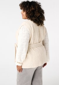 Beige gewatteerde vest met een riem in de taille, gelaagd over een witte textuur long-sleeve top. Grijze high-waisted broek maakt de outfit compleet.