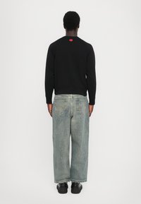 Zwarte sweatshirt met rode logo-detail, gecombineerd met lichtblauwe loose-fit jeans en zwarte sneakers, gezien vanuit de achterkant.