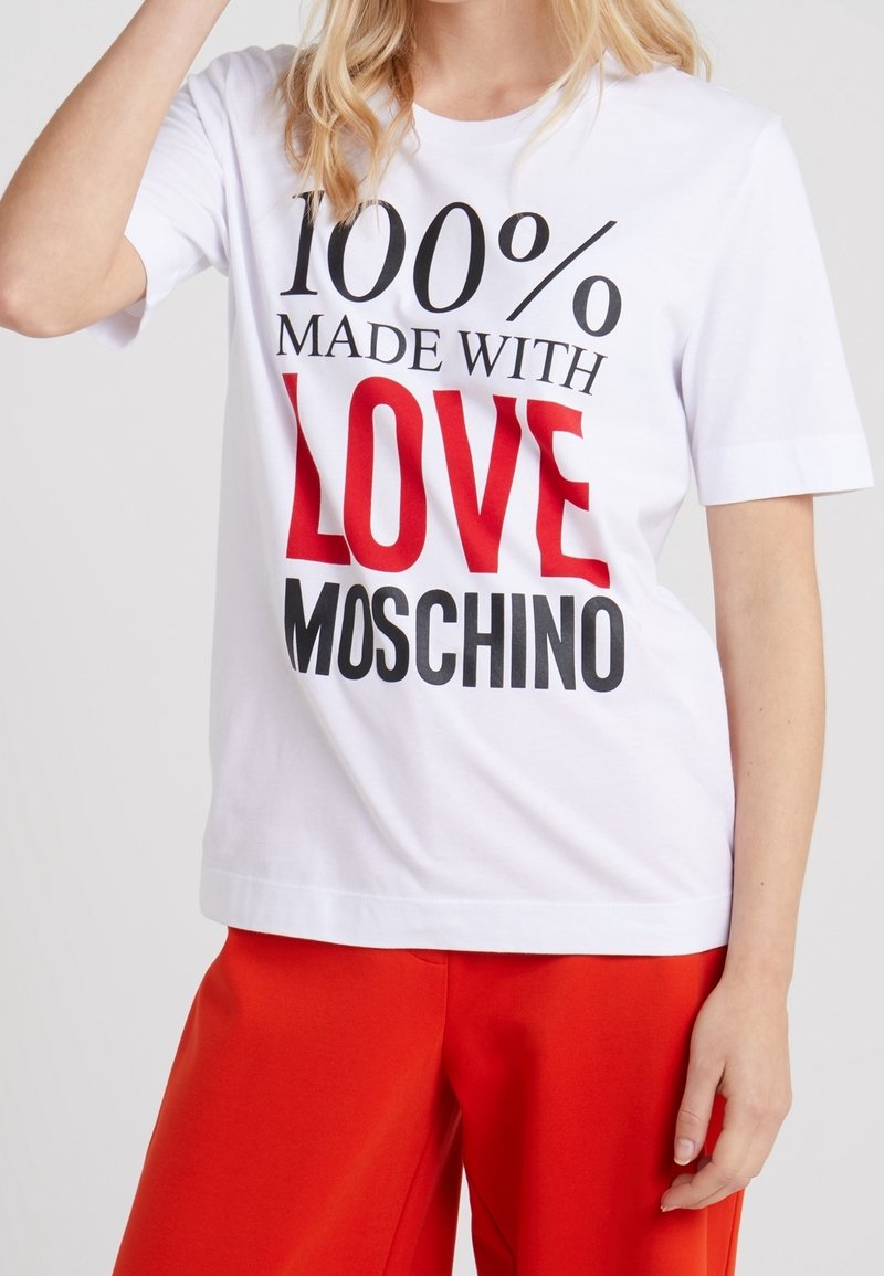 Bílý bavlněný tričko s krátkým rukávem, zdobené tučným černým a červeným textem "100% VYROBENÉ S LÁSKOU MOSCHINO." Uvolněný střih.