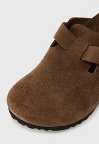 Καφέ σουέντ παπούτσι slip-on με στρογγυλεμένο μπροστινό μέρος, με δύο κλεισίματα με λουράκια και υφή στην σόλα. Ομαλή επιφάνεια με απαλή, βελούδινη  αίσθηση.