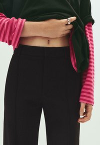 Černé tailored kalhoty v kombinaci s růžově pruhovanou košilí s dlouhým rukávem. Košile má vyhrnuté rukávy a volný střih, který odhaluje břicho.